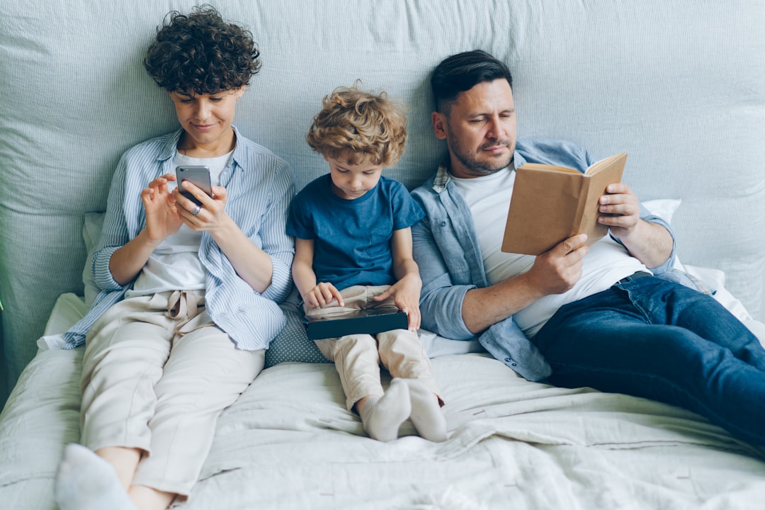 Mother using smartphone son holding tablet father reading book in bed at home by Best Kids Tablets a man and two children are sitting on a bed
