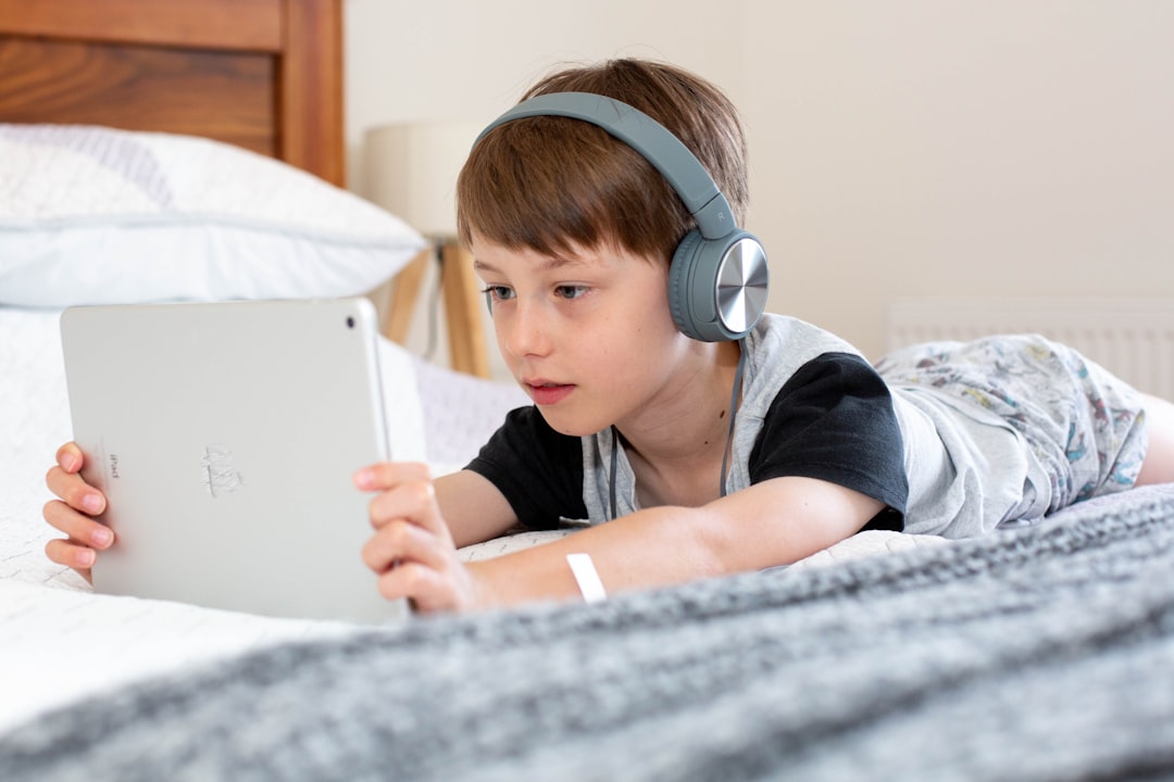 Boy using iPad with headphones on by Best Kids Tablets boy in blue shirt wearing headphones lying on bed